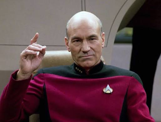 Patrick Stewart as Captain Picard in a red Starfleet uniform, seated on the bridge and raising one finger.