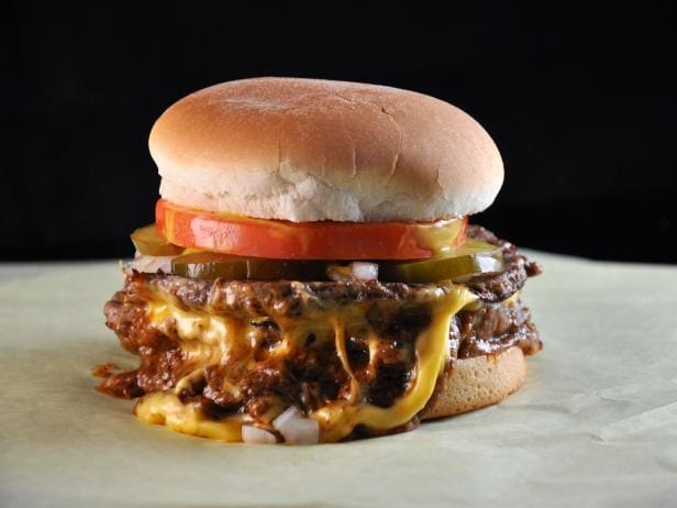 A Tommy's chili cheeseburger on paper: a toasted bun over a slice of red tomato, pickle chips and chopped onion, with dark beef chili and melted yellow cheese running off the patty.