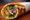 A Kolkata kathi roll: spiced chicken with sliced red onion and chopped coriander chutney in a charred, flaky paratha, half-unwrapped from white paper.