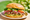A shredded-pork sandwich on a sesame roll on a paper plate outdoors, pulled pork under shredded lettuce, tomato, pickled red onion, avocado and coriander against blurred greenery.