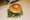 A grilled bagel sandwich stuffed with spinach and sprouts on deli paper, prep trays behind it