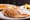 Pancakes on a white black-rimmed plate with syrup pouring over them, scrambled eggs on a plate behind