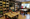Deli interior: wooden tables with red metal chairs in front of shelves of jarred and packaged goods, window at right