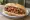Choripán con Salsa Criolla: an illustration of pork, sausage, tomato, onion and more on baguette.