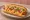 Hot Dog con Queso: an illustration of sausage and cheese on hot dog bun.