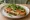Kanapka z Hummusem: an illustration of hummus, cucumber, tomato and arugula on bread.