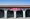 White stucco storefront with arches and a red tile roof under a red Angelo's Gourmet Delicatessen sign, neon Open sign lit.