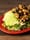 A salad on a red plate with sliced avocado, grilled chicken, black beans, corn and tortilla strips on a wood table.