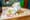 A halved chicken Caesar sandwich in white paper on a bamboo board, shaved parmesan and cucumber scattered around it.