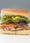 Turkey sandwich on a soft white roll with cheddar, tomato, red onion, lettuce and pepperoncini, on white deli paper.